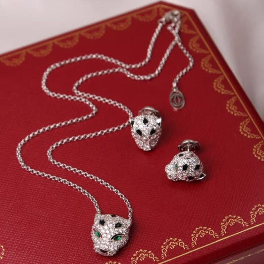 A jewelry set featuring a silver-toned necklace and matching earrings with panther head pendants paved with clear stones and accented with green gemstones, displayed on a red luxury-style jewelry box.