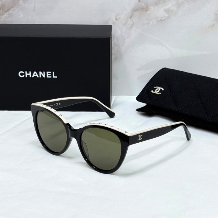 Chanel sunglasses with a black acetate frame, gold metal trim, and dark green lenses, displayed with a black Chanel box, a black quilted pouch, and a white marble background.