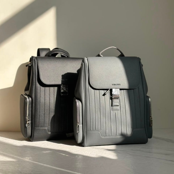 Two dark gray structured backpacks with vertical pleat detailing, displayed side-by-side on a light gray marble surface with dramatic diagonal sunlight casting shadows.