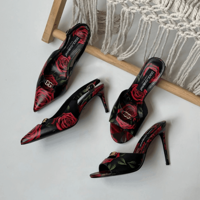 Four black high-heeled mule sandals with red rose prints and gold-tone hardware, arranged on a light surface beside a woven rope chair.